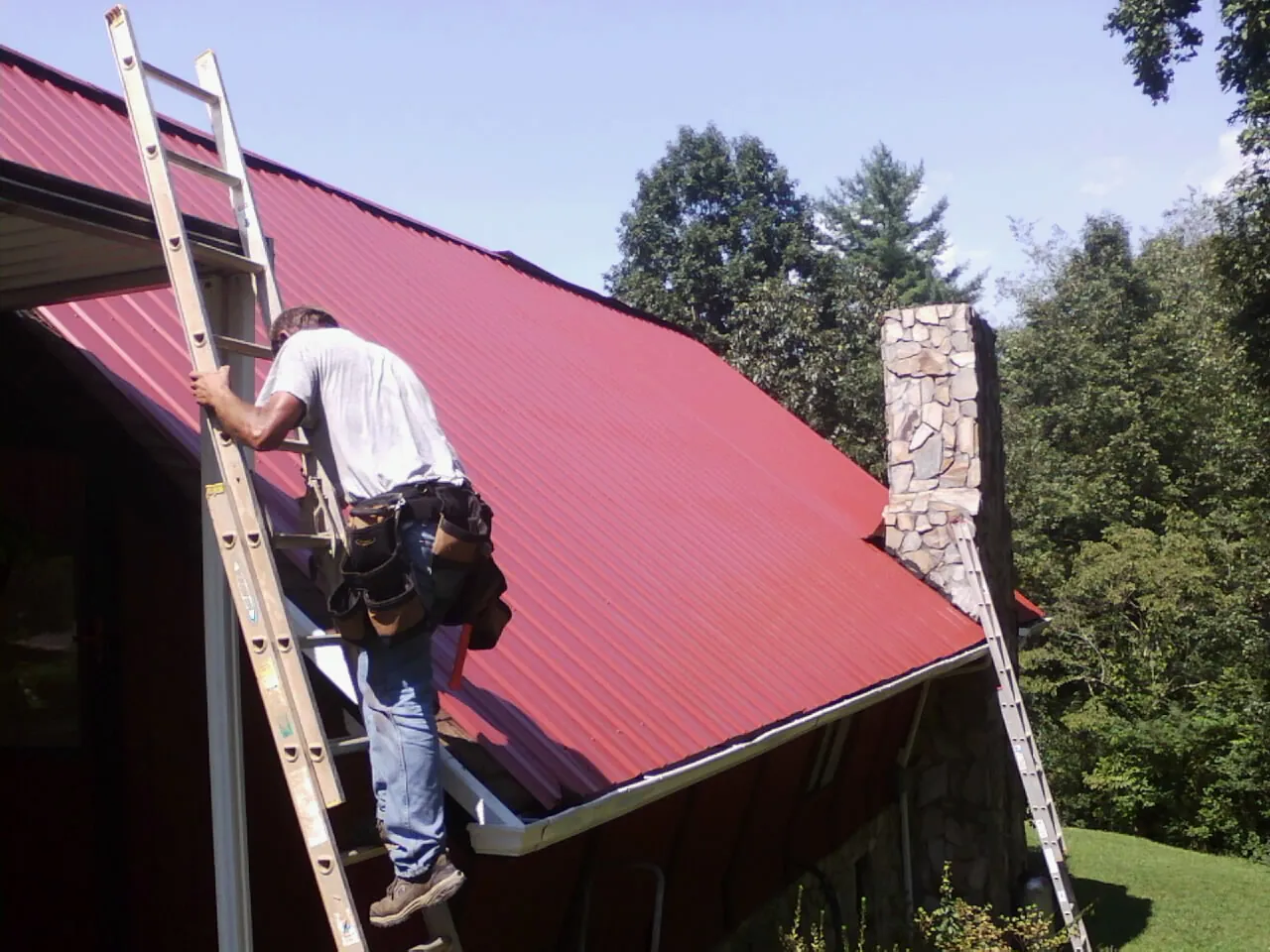 Roofer on a ladder beside a red metal roof and stone chimney — Soffit & Fascia Repair by Roofing Brothers in Jelm